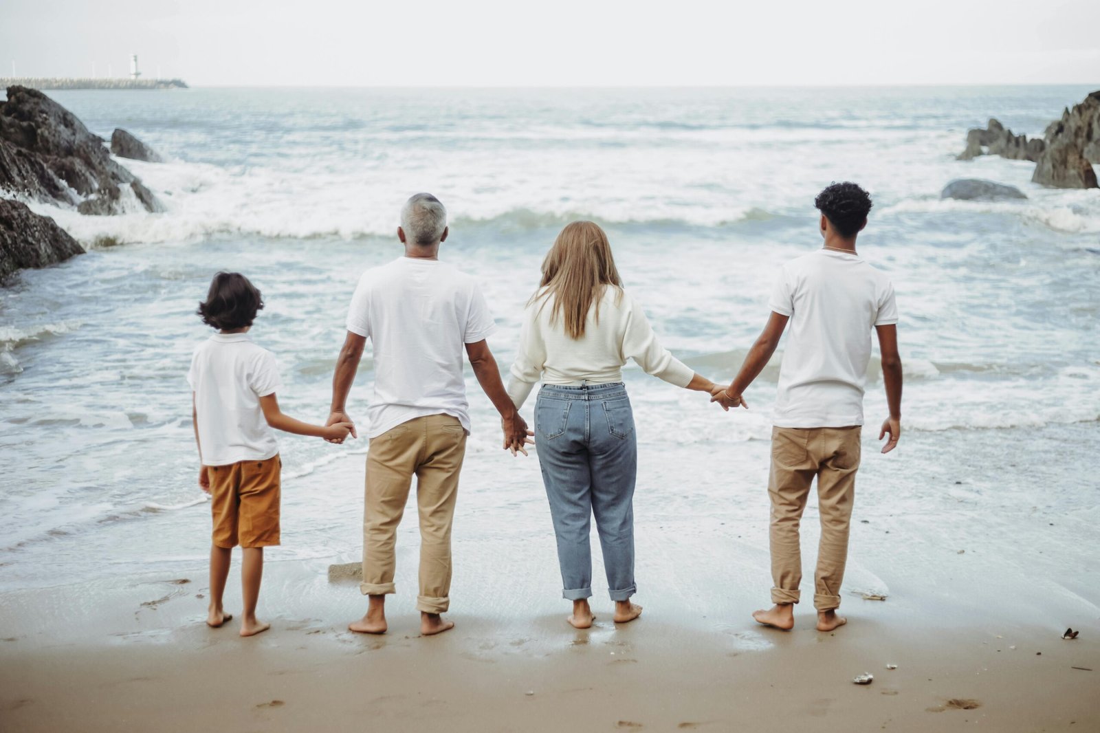 Tourism A family of four holds hands while facing the ocean, enjoying a peaceful day at the beach.